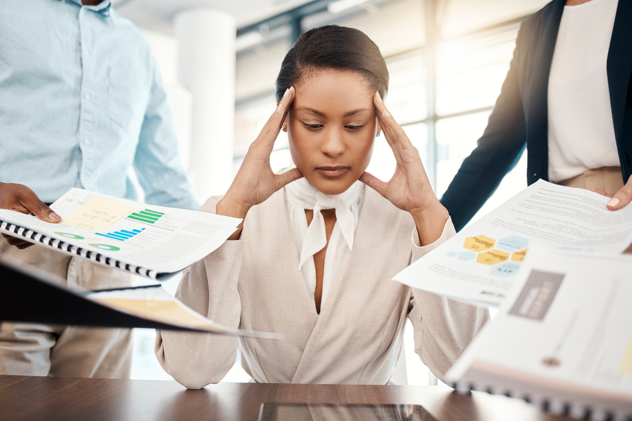 Professional woman sitting at a desk holding her head and experiencing anxiety while colleagues hold papers in front of her. 